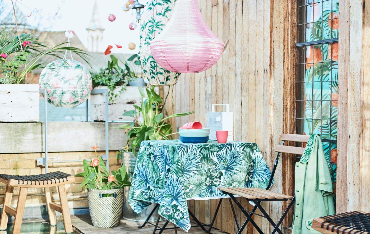 A city roof terrace with a patio table covered with leaf-print fabric, folding chairs, potted plants and coloured lanterns.