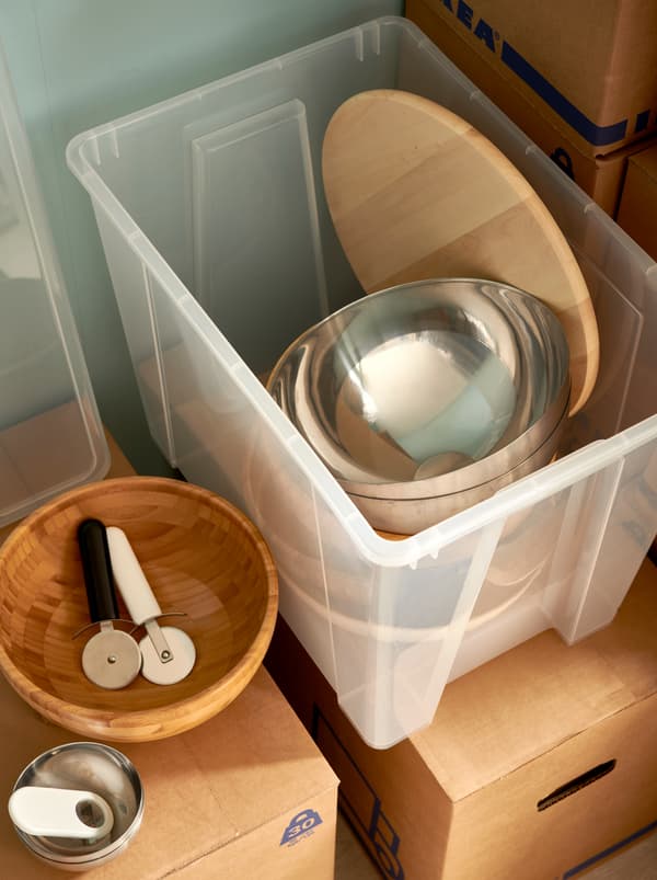 A clear-plastic SAMLA box, filled with BLANDA serving bowls, a lazy Susan and tableware, all placed on stacked JÄTTENE boxes.