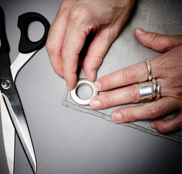 A close-up image of hands, cutting two holes then pushing the eyelets into them on grey fabric.