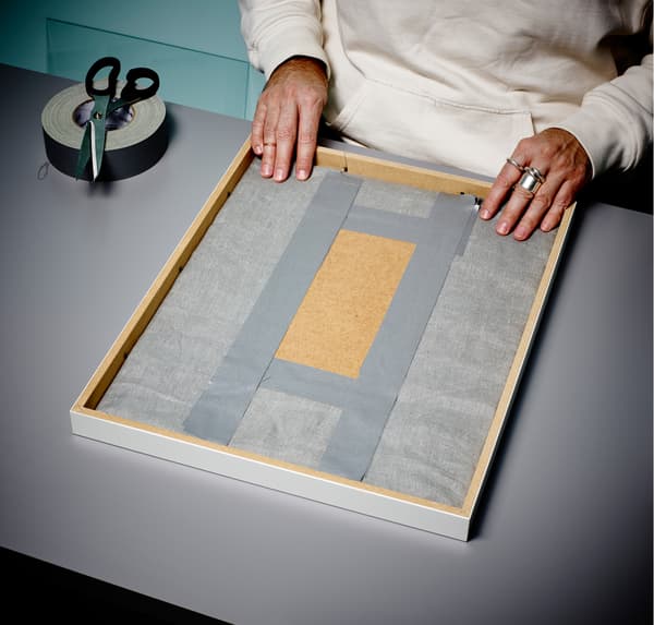 A close up of a person’s hands, placing a grey fabric-covered back of a frame into a photo frame, and folds over clips.