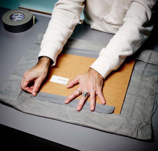 A close up of a person’s hands, tapes grey fabric to the back of a photo frame.