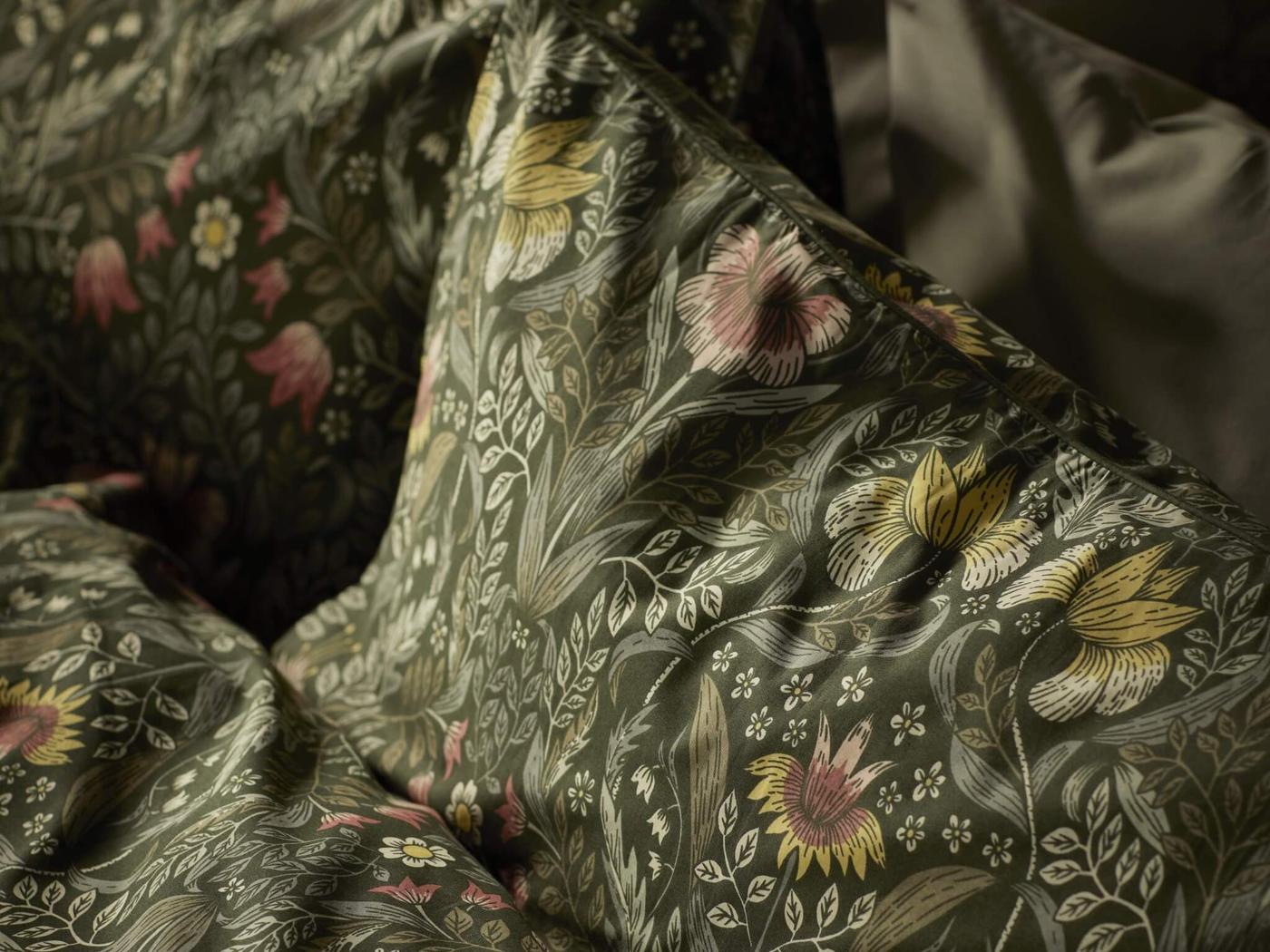 A close-up of bedding with a detailed floral pattern. The pattern features various flowers and leaves in shades of pink, yellow, and green on a dark background.