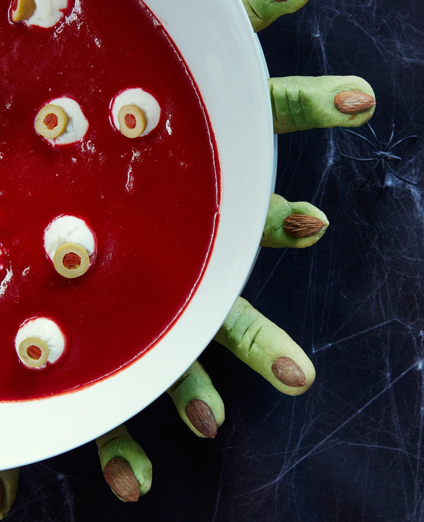 A close up of Halloween food resembling blood with eyeballs floating in a white bowl with green cookies resembling fingers displayed around it.