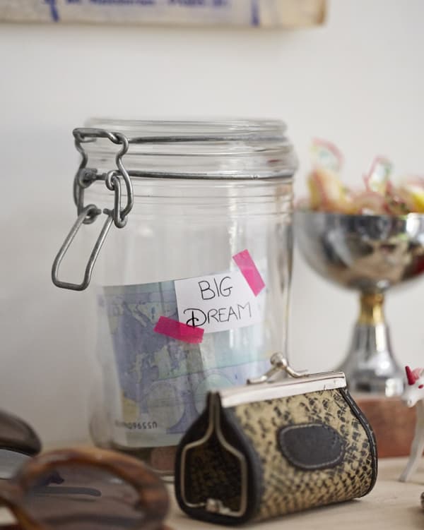 A closeup of a clear glass jar with money and bills inside with a taped note that reads 'Big Dreams' to save up for a goal.