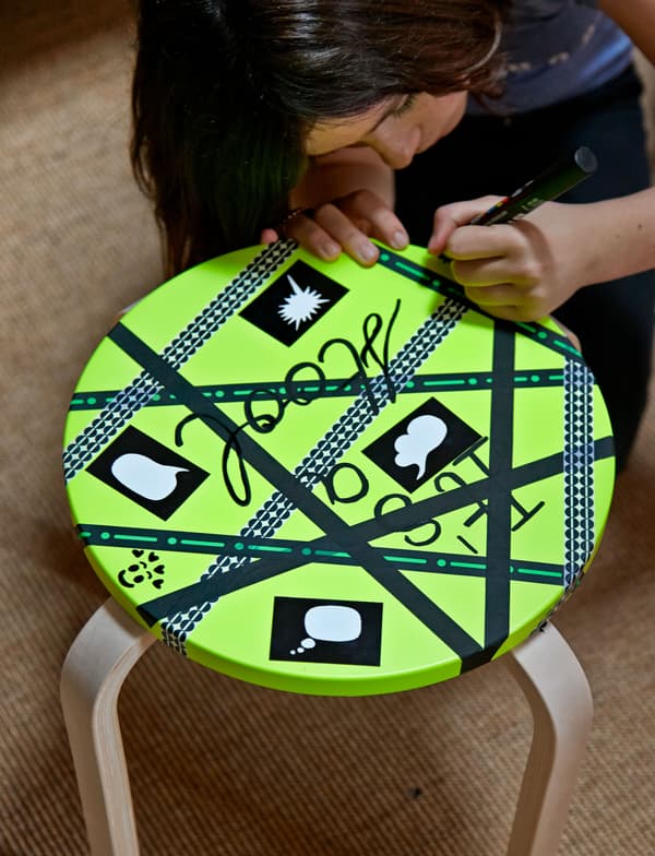 A closeup of a stool's seat painted lime green and being decorated with black deco tape and handwritten words.