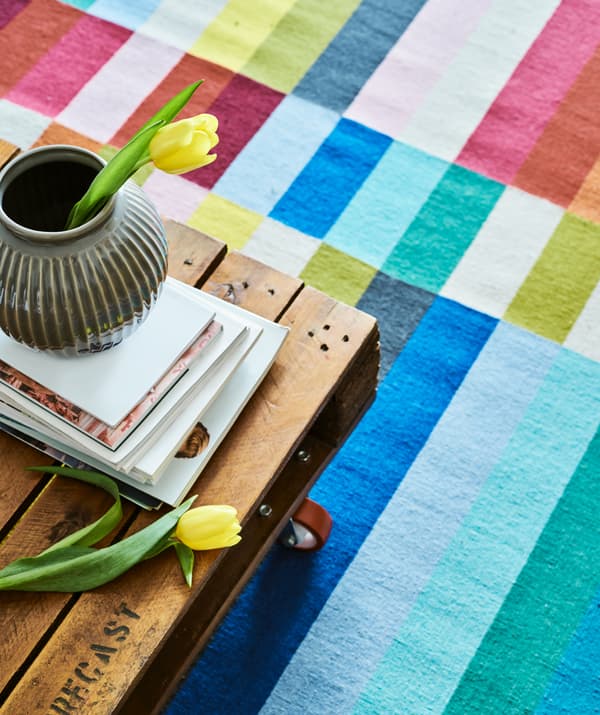 A coffee table made from a repurposed palette and red castors on a colourful stripy rug, magazines and a grey vase on top.
