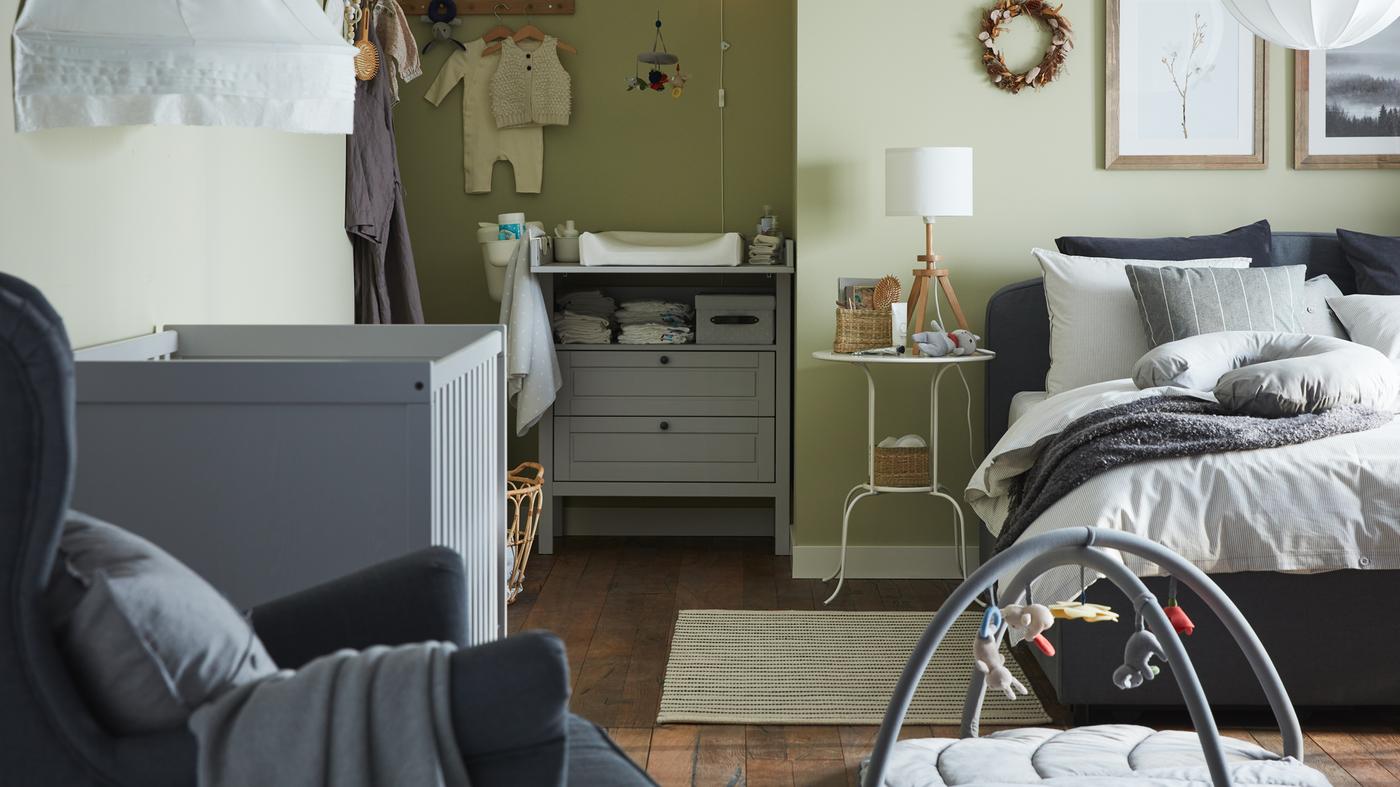 A combined bedroom and baby room with a grey upholstered bed, grey cot, grey changing table/chest of drawers, green walls.