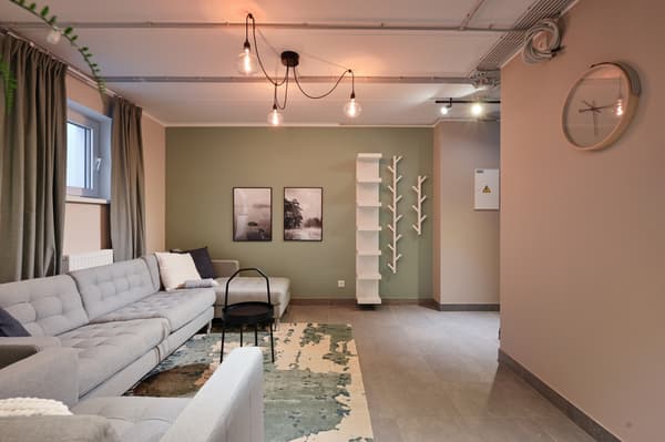 A contemporary co-working space with a lounge area featuring a light grey sofa, soft lighting, indoor plants, and muted green walls. Open shelving, minimalist furniture, and warm neutral tones create a calm and inviting atmosphere for work, meetings, and relaxation.