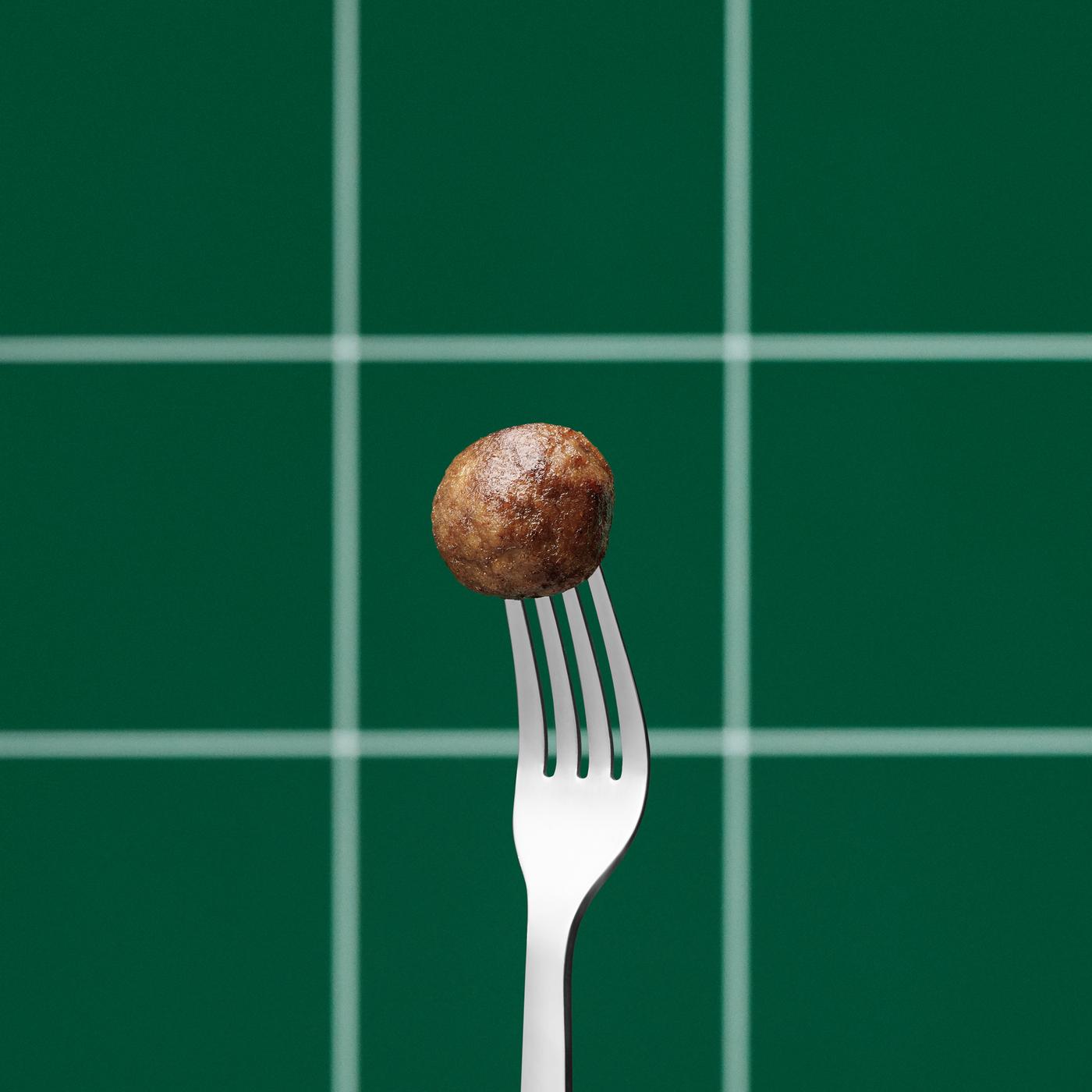 A cooked IKEA plantball on the end of an upright silver fork, against a green and white, check pattern background.