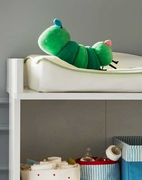 A corner of a baby changing mat with KLAPPA caterpillar soft toy on it and baby milk bottles and cloths in baskets on the shelf below.