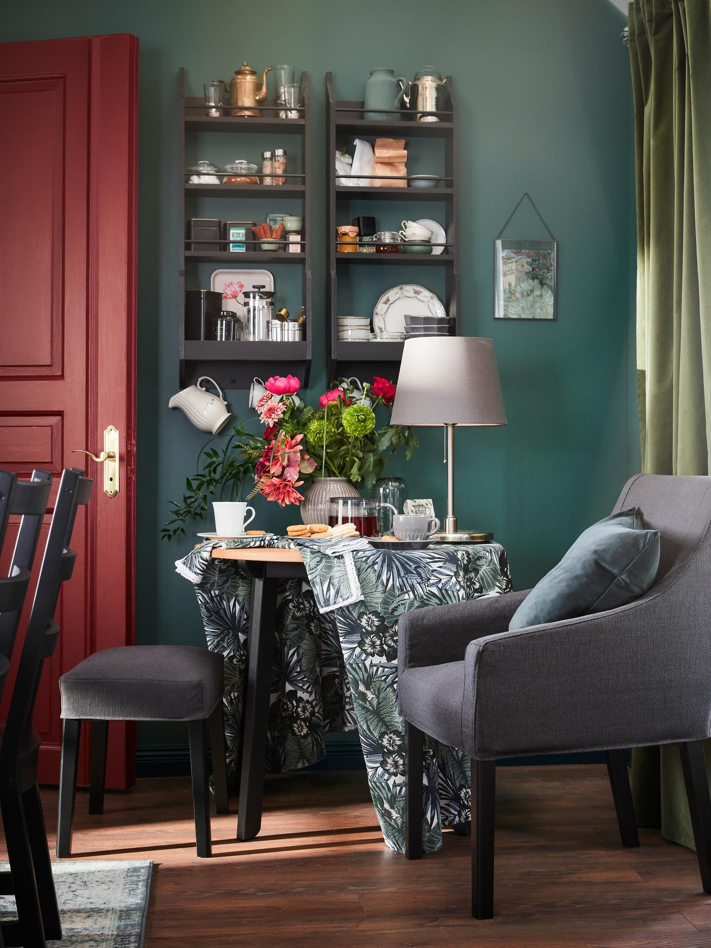 A corner with a black/dark grey chair with armrests, a table where tea is served and a window with white/green curtains.