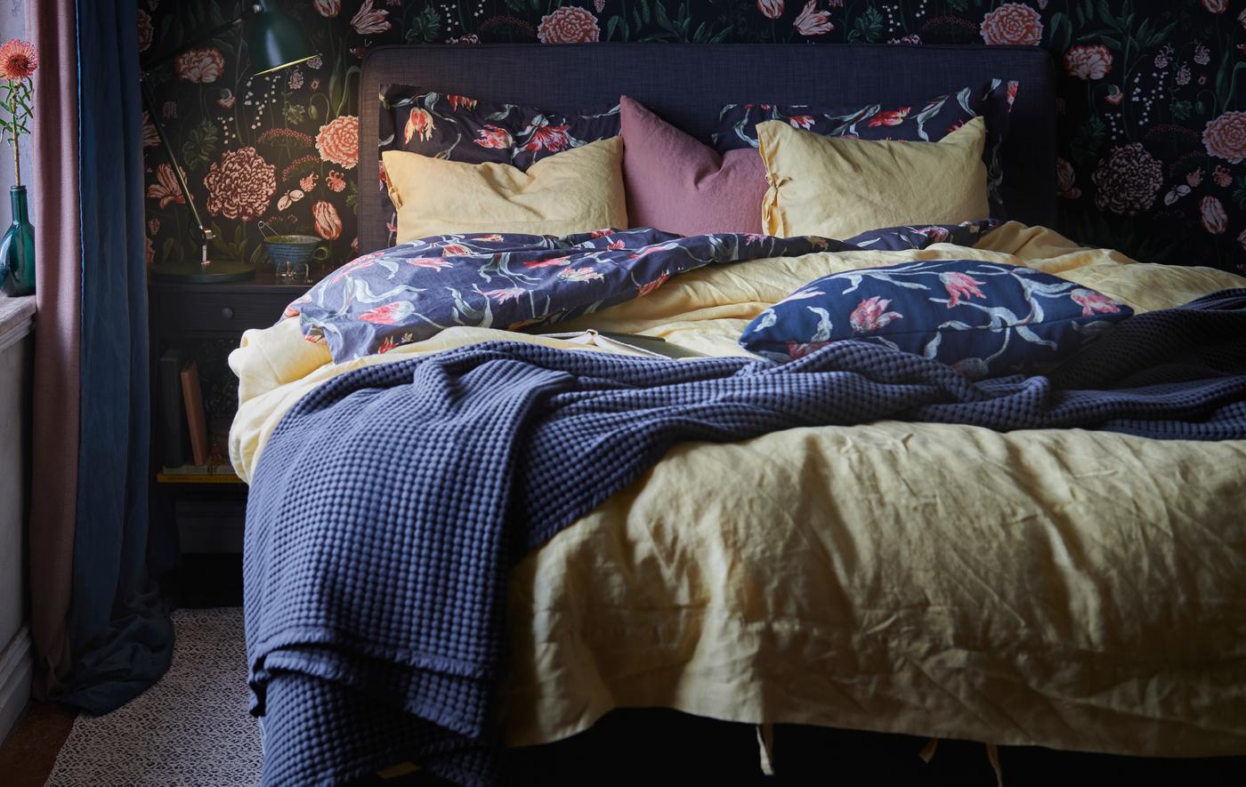A cosy bed with two duvets, pillows and a bedspread. Next to it is a bedside table with a lit lamp and a mug.