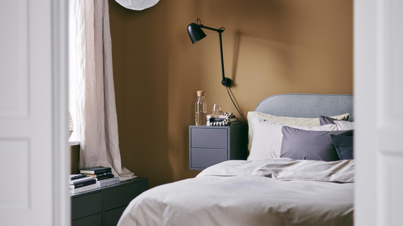 A cosy bedroom with rich caramel walls and a VADHEIM upholstered bed frame, a wall lamp and a bedside table.