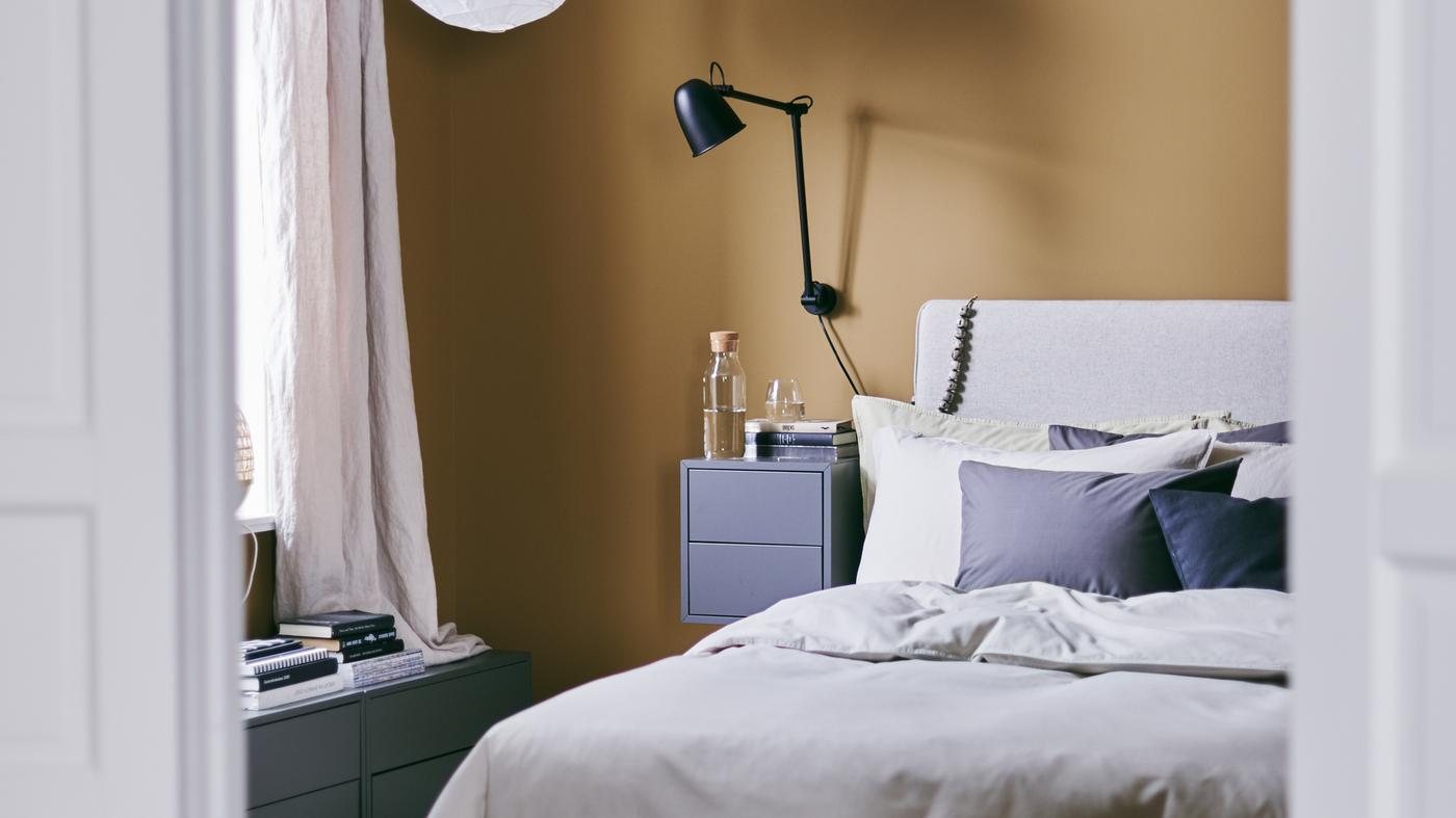 A cosy bedroom with rich caramel walls and the DUNVIK bed frame, a wall lamp and a bedside table.