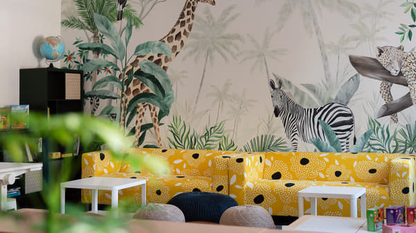 A cosy seating area with bright yellow patterned sofas placed in front of a tropical jungle mural featuring animals including a giraffe, zebra, leopard, and toucan. Small white tables and soft seating create a playful and welcoming atmosphere.