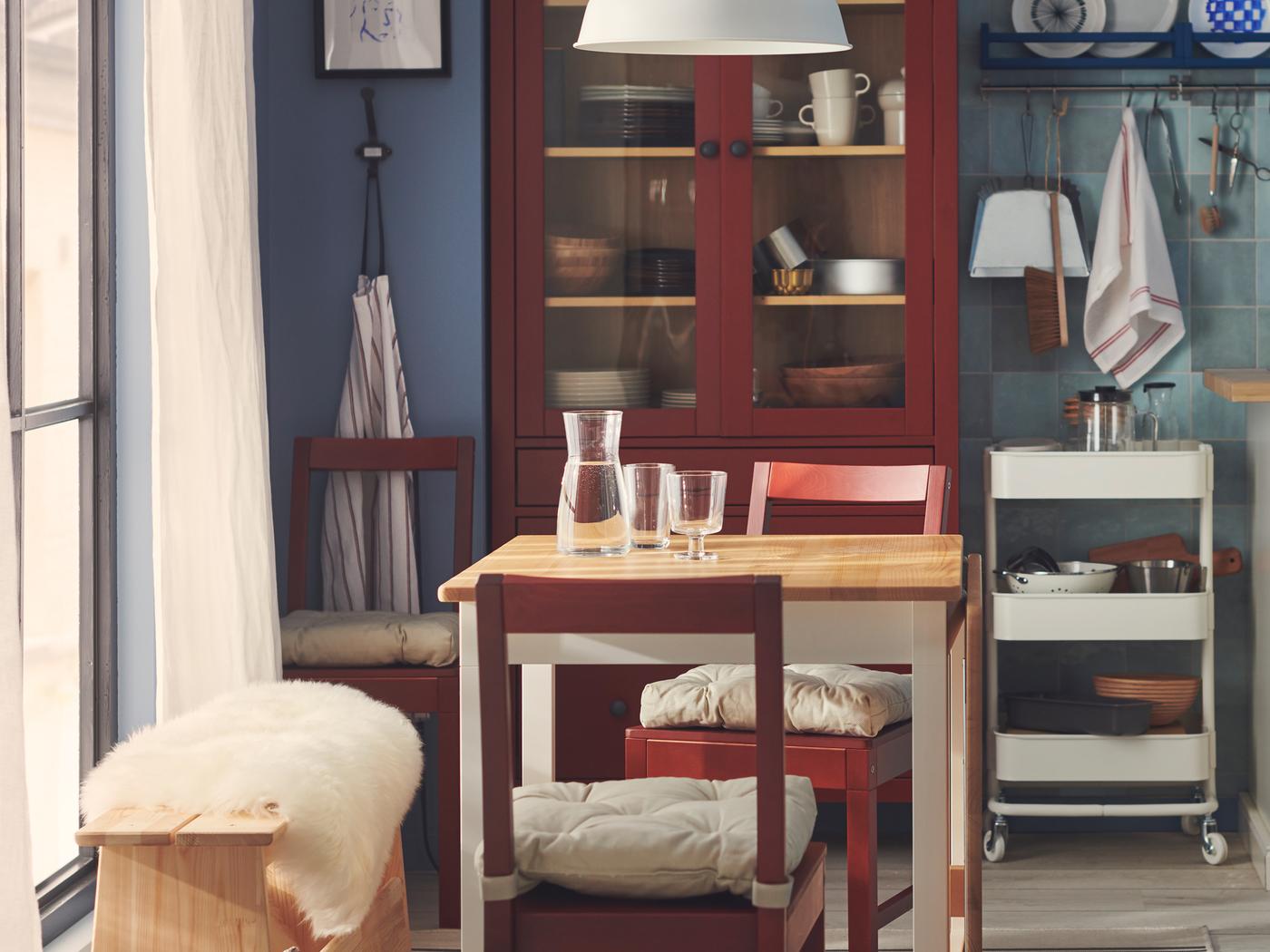 À côté d’une grande fenêtre se trouve une petite table pour deux avec des pieds blancs et un plateau en bois brun clair ainsi que deux chaises rouges.