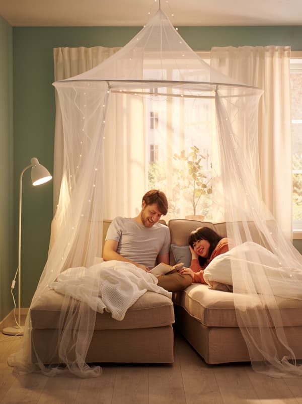 A couple lying under VÅRELD bedspreads in two side-by-side chaises longues by a sunlit window, a SOLIG net draped over them.
