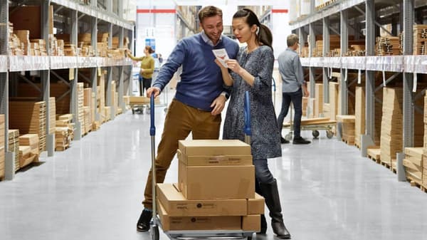 A couple shopping in an IKEA warehouse pushing a flat cart with boxes on top