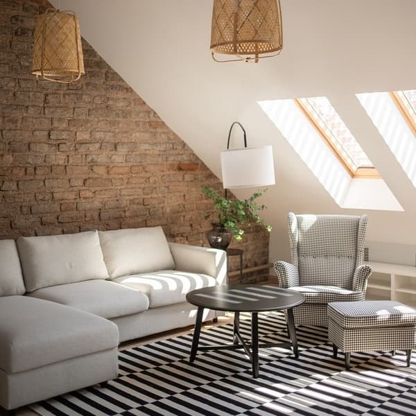 A cozy attic living room featuring a sloped ceiling with skylight windows that let in abundant natural light. The space combines rustic and modern elements, with an exposed brick wall, a light grey sectional sofa, a black-and-white checkered armchair and footstool, a black round coffee table, and a striped rug. Woven pendant lights and green plants add a touch of warmth and texture.