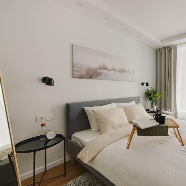 A cozy modern bedroom featuring a grey upholstered bed with soft cream bedding and layered pillows. On the bed is a wooden breakfast tray holding a book, cup, and wine glass. Minimalist black wall sconces frame a nature-themed canvas above the bed, and the space is softened with full-length curtains, potted greenery, and small round bedside tables.
