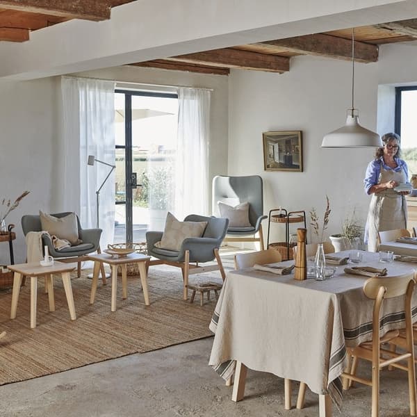 A cozy, sunlit dining and living room with rustic charm. The space features light wood furniture, including a dining table set for four with a beige tablecloth and wooden chairs. In the background, a sitting area includes two blue-gray armchairs and a matching high-back lounge chair, all with neutral cushions. A woven rug and several small wooden coffee tables create a warm, inviting feel. Exposed wooden ceiling beams and concrete flooring add rustic character, while a large sliding glass door lets in natural light. A woman stands in the background near the kitchen area, wearing a blue shirt and apron.