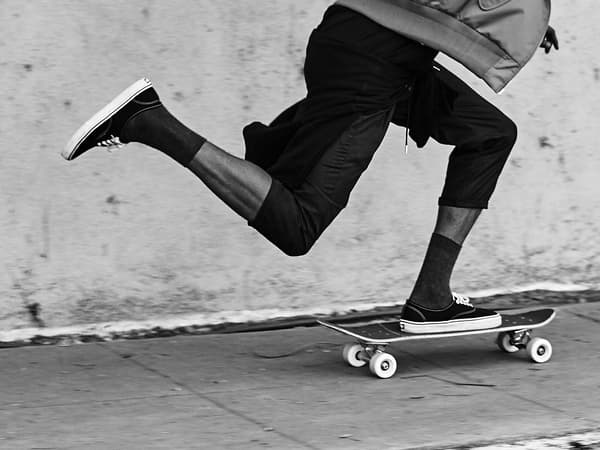 A cropped black and white image showing a young man's legs skateboarding up a sidewalk.