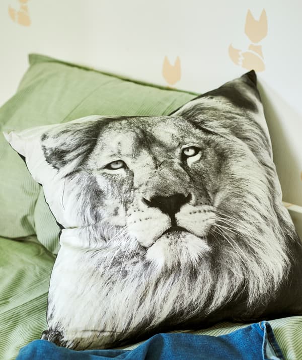 A cushion with a black and white photo of a lion on a bed with green bedding, the wall is stencilled with a fox pattern.