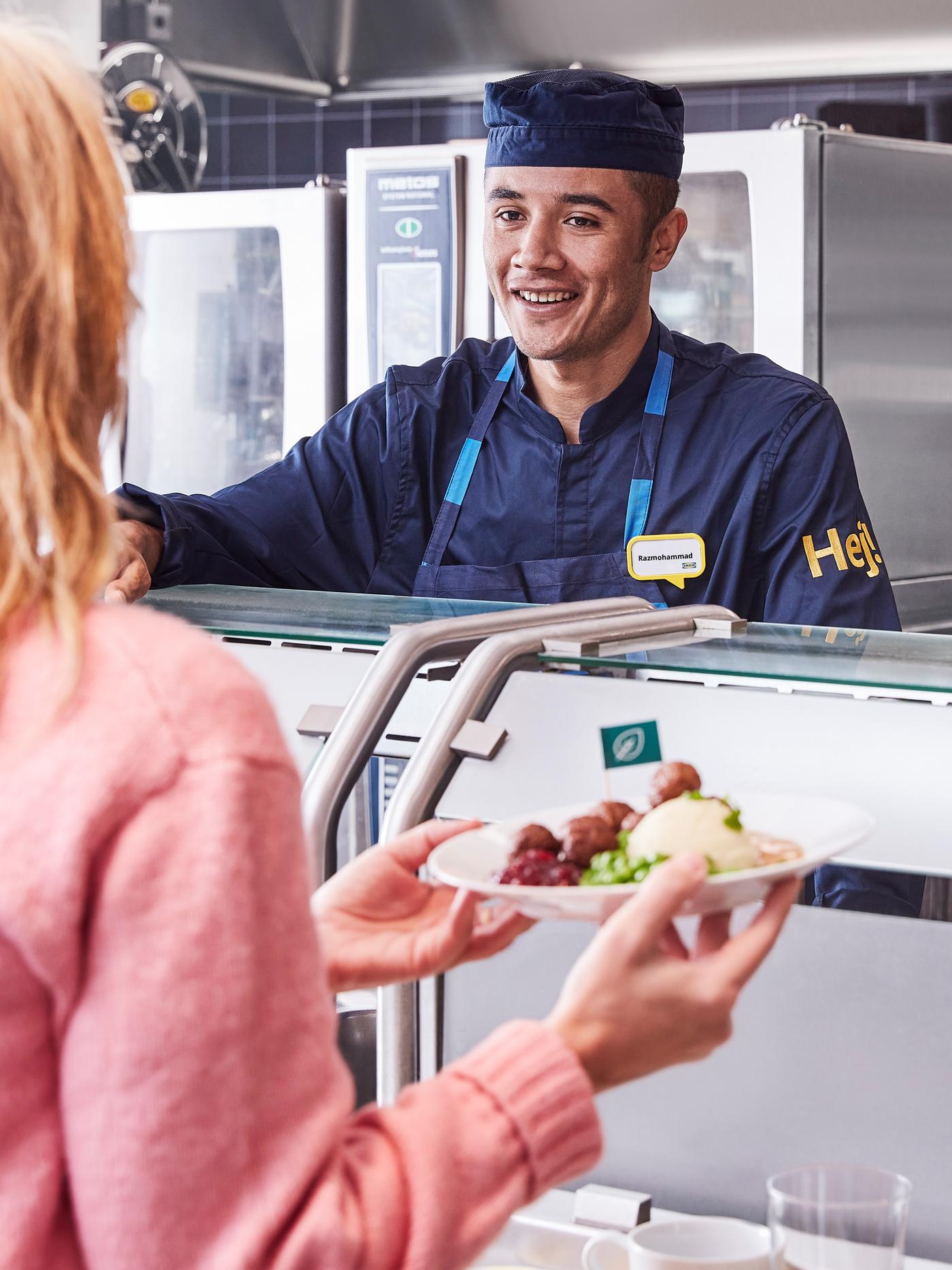 A customer wearing a pink jumper accepts her meal from a smiling Swedish Bistro co-worker wearing a dark blue uniform.