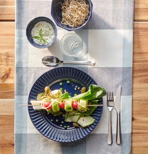 A dark blue plate with colourful vegetables on a skewer, MOPSIG cutlery and a glass of water on a blue chequered table cloth.