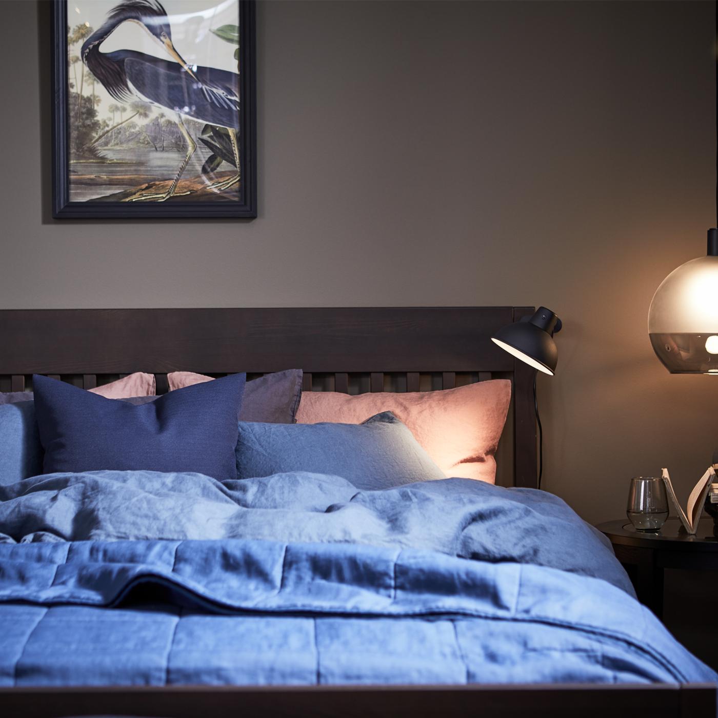 A dark blue PUDERVIVA duvet cover on an IDANÄS bed frame with storage with a lit JAKOBSBYN pendant lampshade to the right.