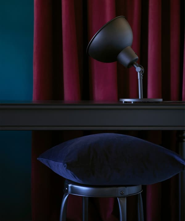 A dark blue velvet cushion perched on a stool, in front of a black wood desk and dark red curtain.