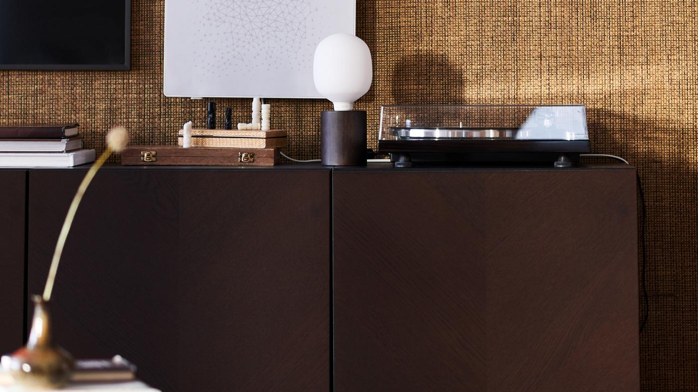 A dark-brown BESTÅ cabinet against a hessian wall, a BLEKKLINT table lamp and a SYMFONISK picture frame with WiFi speaker.