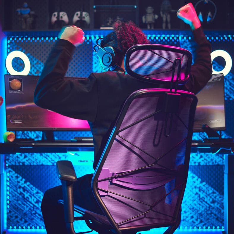 A dark gaming room with a lit-up gaming station. A person, sitting in a gaming chair, is raising the arms victoriously.