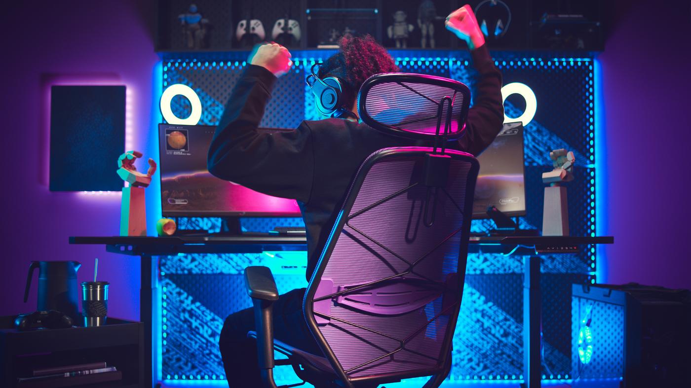 A dark gaming room with a lit-up gaming station. A person, sitting in a gaming chair, is raising their arms victoriously.
