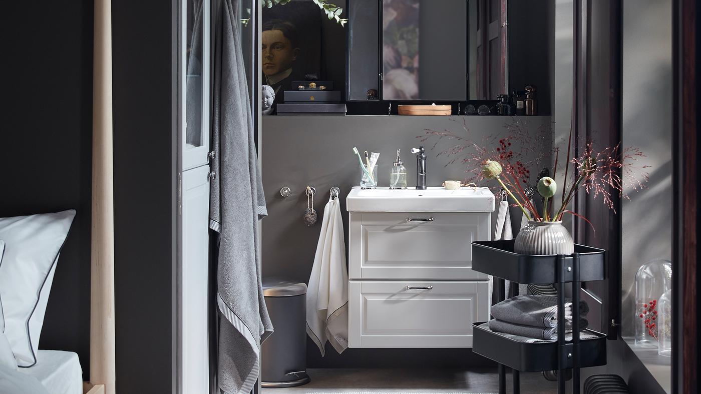 A dark grey bathroom with a wash-stand in light grey, a black RÅSKOG trolley with folded towels and decorative flowers.