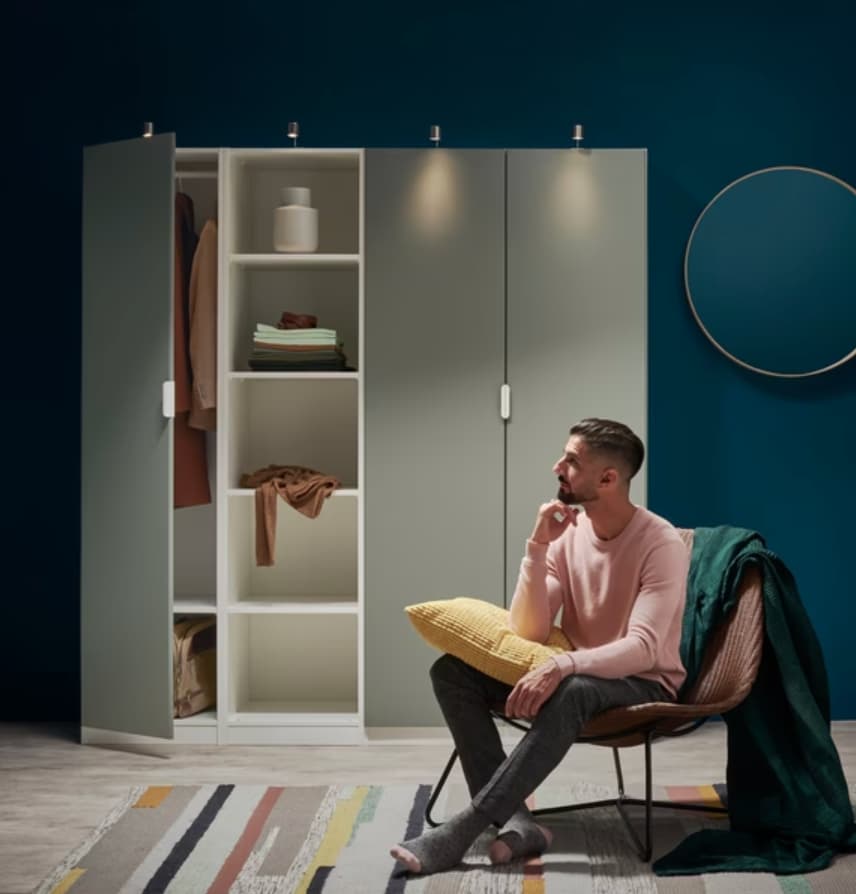 A dark-haired man in a pink shirt sitting and there is an open PAX wardrobe combination behind him.