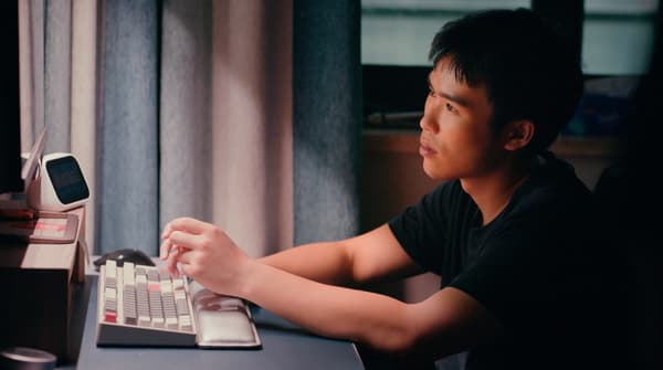 A dark-haired young man is sitting in front of a keyboard watching a computer screen in ambient lighting.