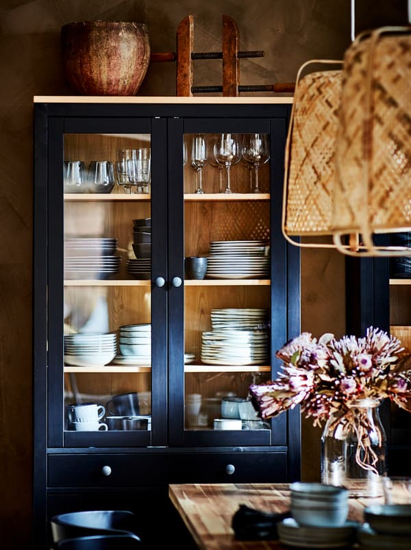 A dark HEMNES glass-door cabinet filled with stacks of tableware, standing by a wall next to a dining table.
