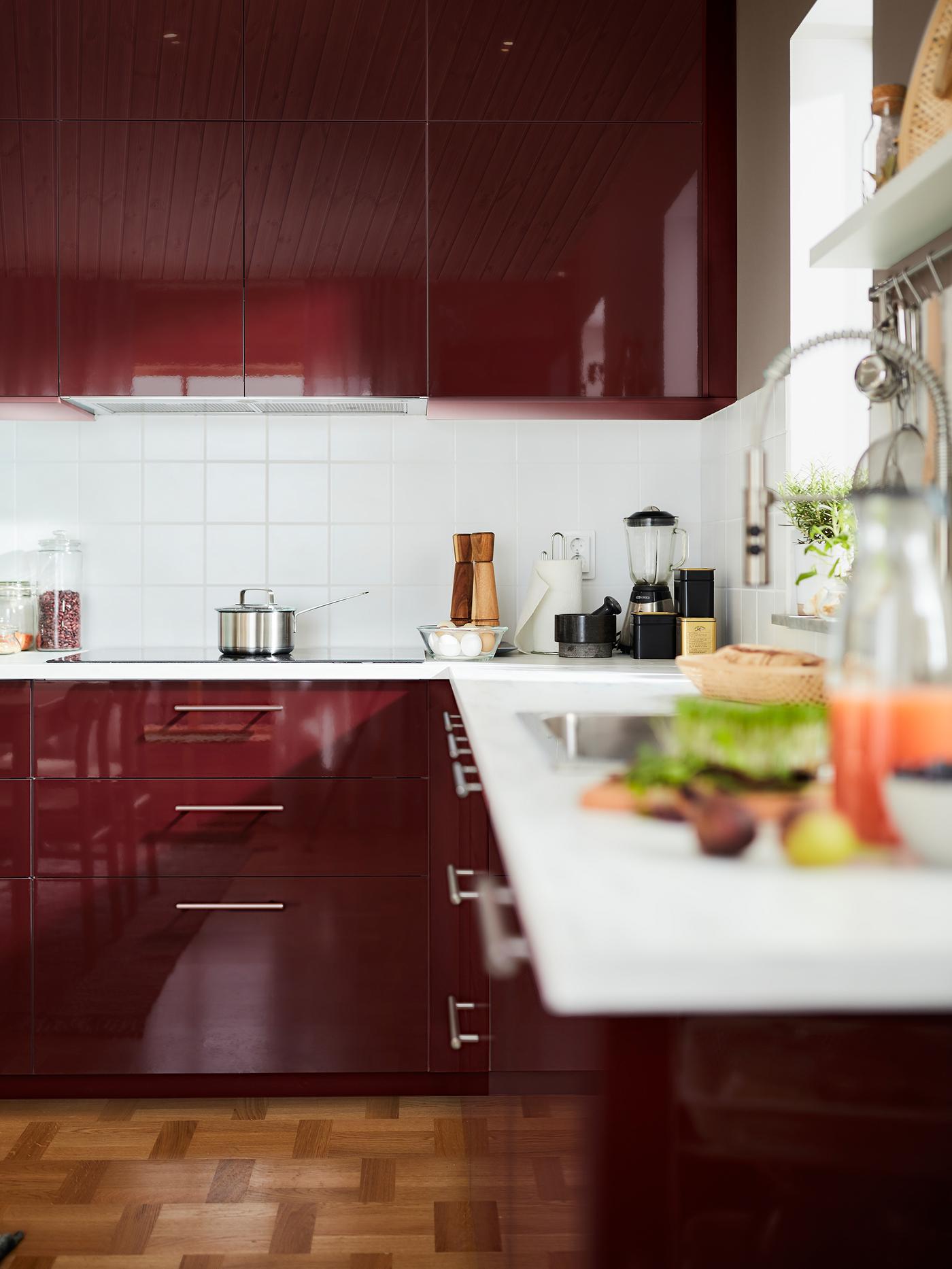 A dark red-brown high-gloss kitchen with a white worktop and a hob. Various kitchen essentials are stored on the worktop.