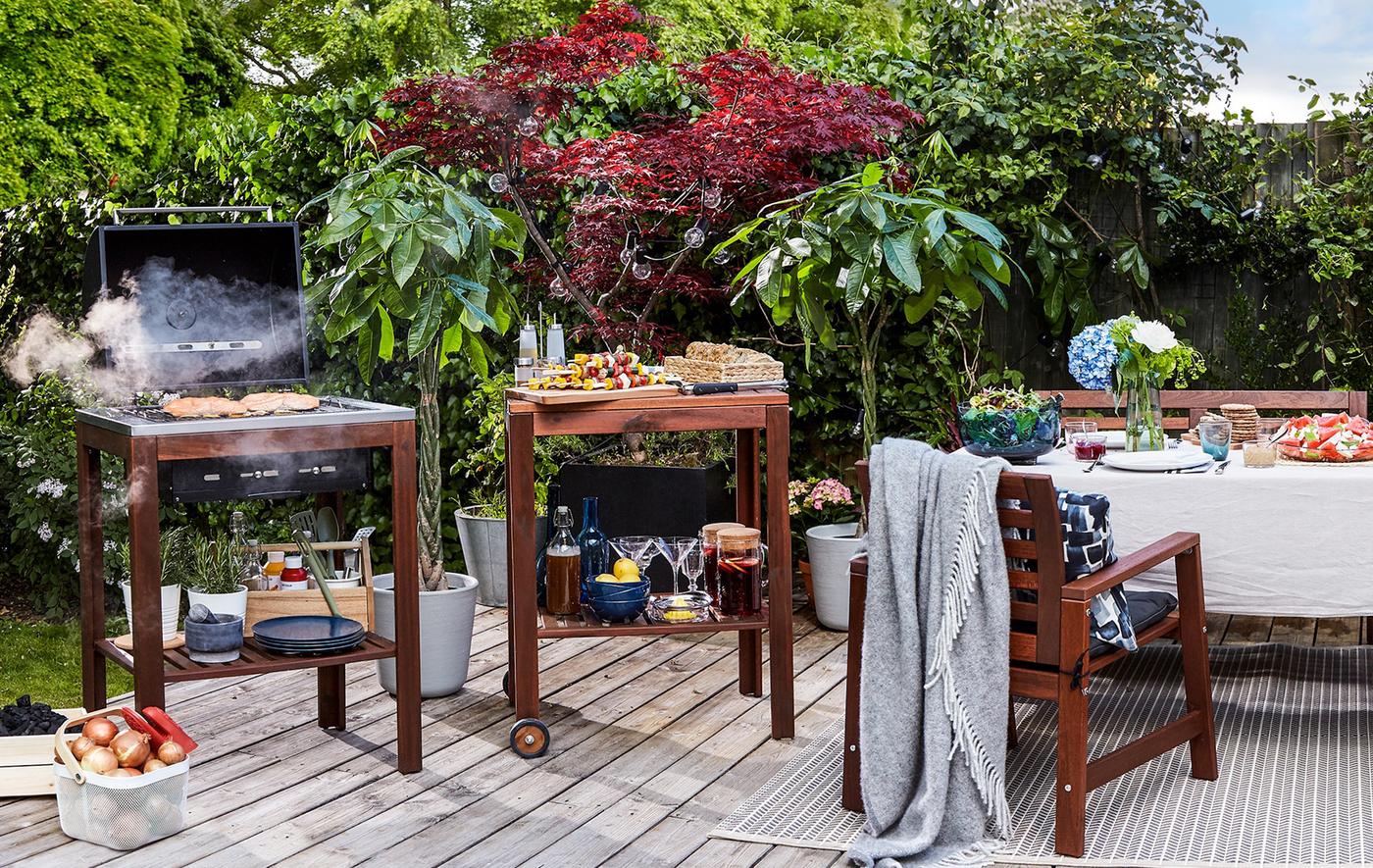 A decked area in a garden with dining table and chairs, food trolley and barbeque.
