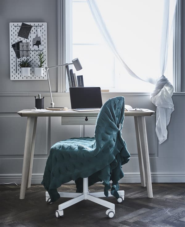 A desk chair covered in a green blanket in front of a minimal wooden desk by a window.