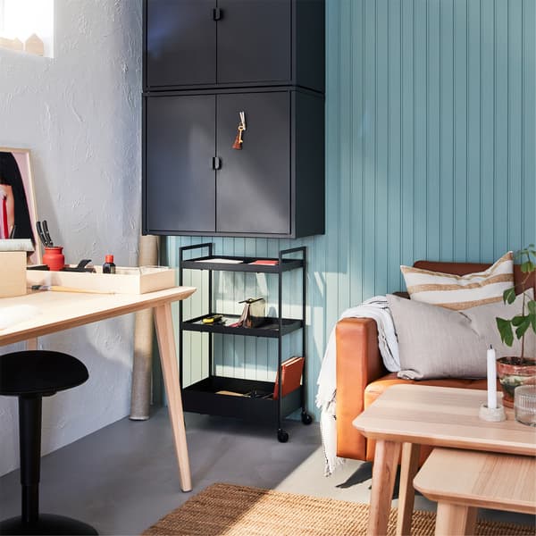 A desk in ash veneer, a jute rug, black wall-mounted cabinets, a black trolley and a golden-brown leather sofa.