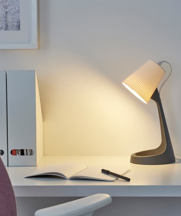 A desk lamp with grey base and white shade and a notepad on a white desk.