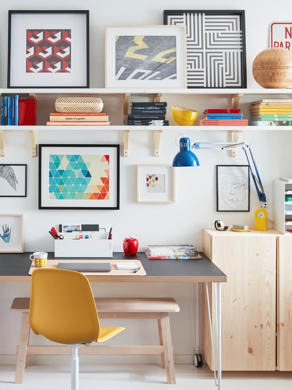 A desk on castors stands by a wall, surrounded by a TERTIAL work lamp, wooden cabinets, wall shelves, and framed artwork.