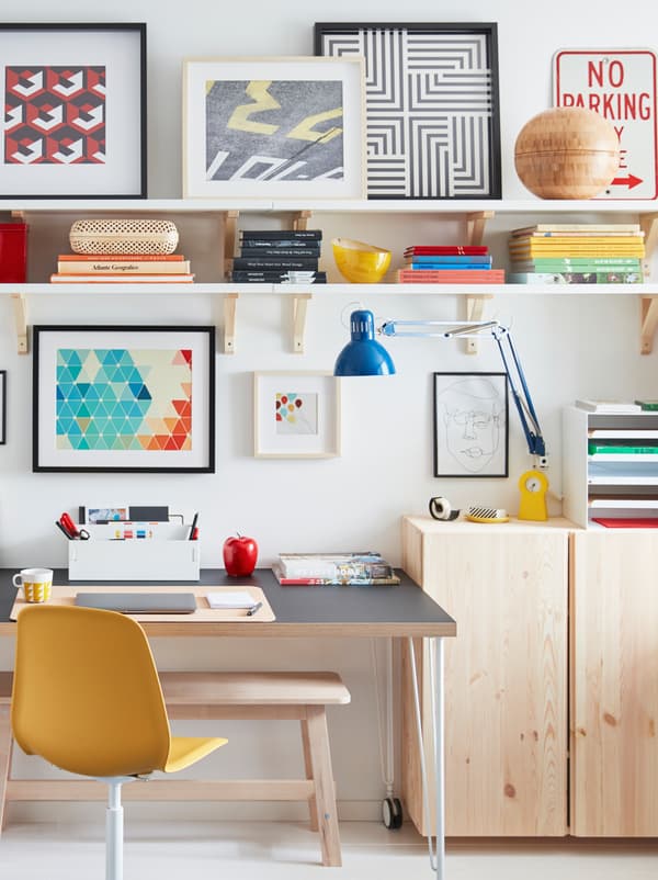 A desk on castors stands by a wall, surrounded by light-wood IVAR cabinets, wall shelves, and framed artwork.