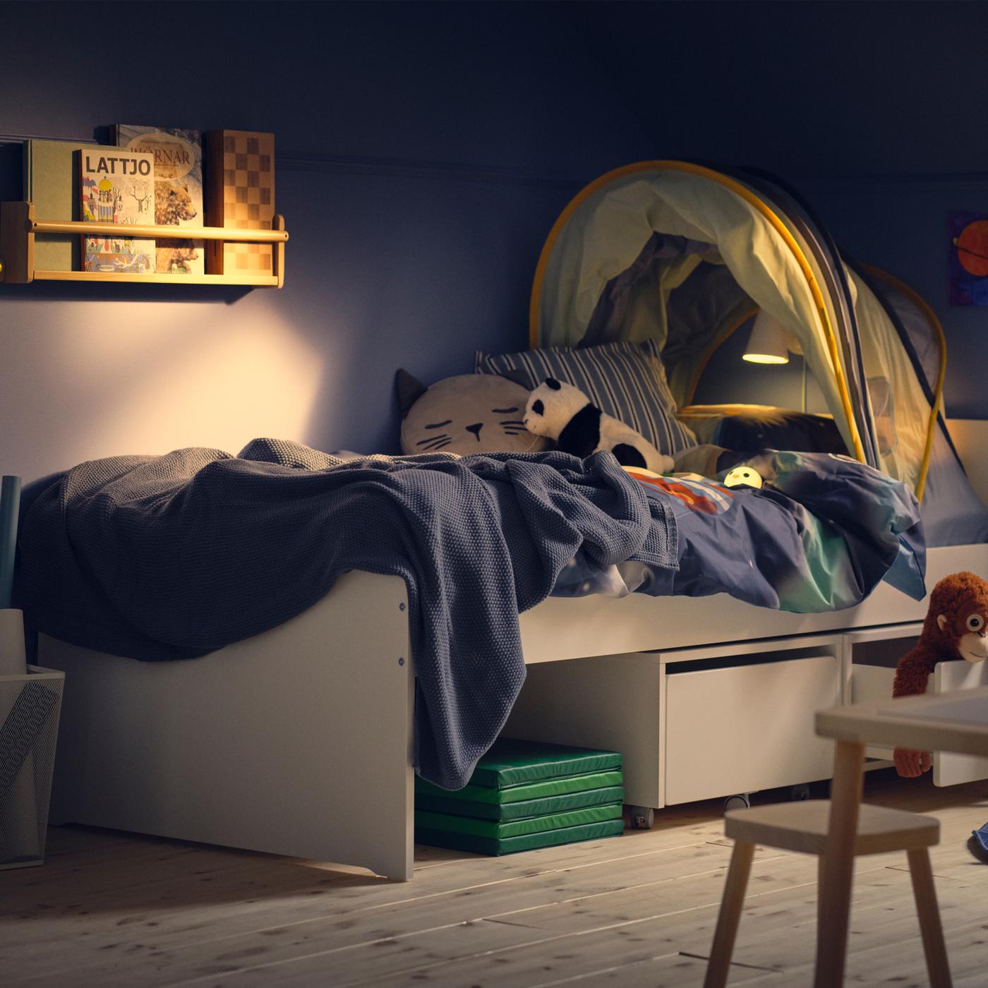A dim blue children’s room with a white SLÄKT bed with storage boxes with castors underneath and an ELDFLUGA bed tent on top.