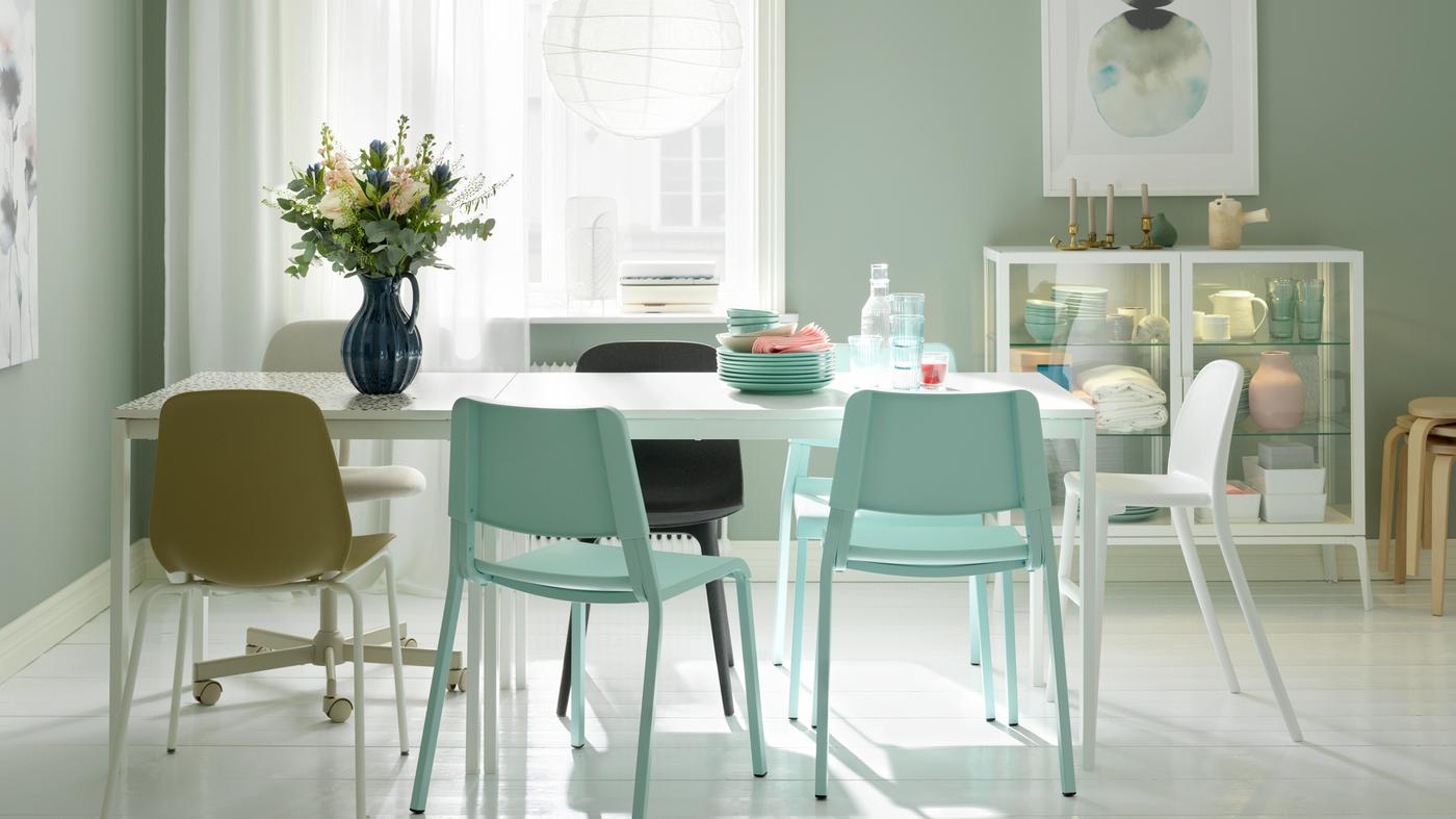 A dining area formed by two white MELLTORP tables in line, a white REGOLIT lamp, assorted chairs and flowers in a vase.
