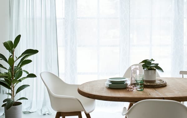 A dining room with a round, wood table and white chairs, in front of French windows dressed with green and white curtains.