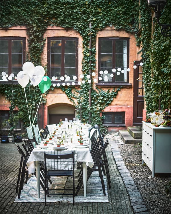 A dining table for a wedding is set up in a courtyard, along with a string of lights, rugs and a chest of drawers.