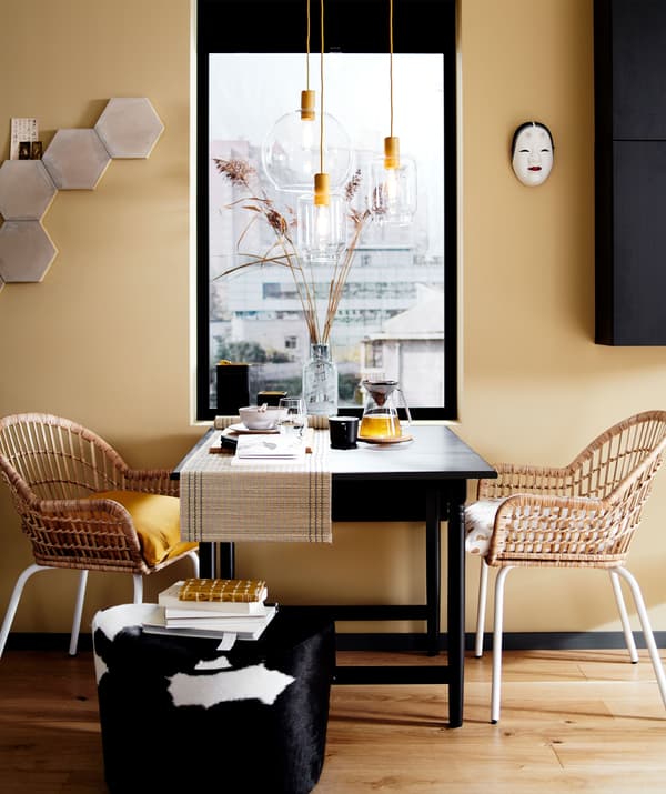 A dining table setup with two rattan chairs and a pouffe in front of a window, plus glass pendant light fittings.